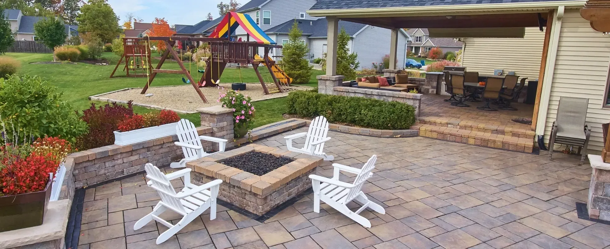 Paver patio with a square stone fire pit and white Adirondack chairs in a backyard